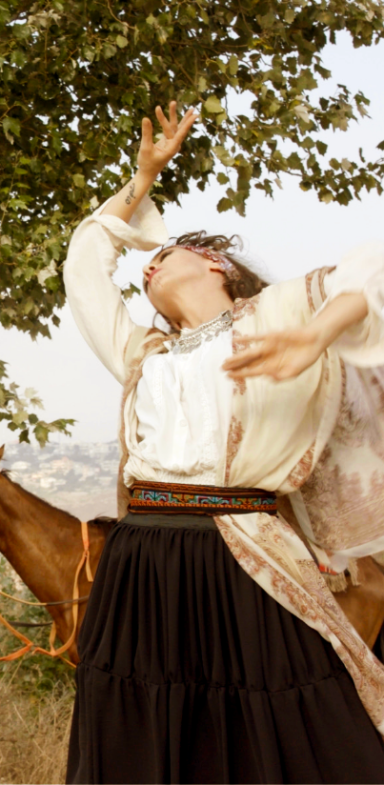 A woman dancing in an open field adorned with traditional clothes and jewelry. Behind her a horse and a tree. 